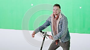 Smiling cute man in a large green background studio he posing in front of the camera while holding his electric scooter