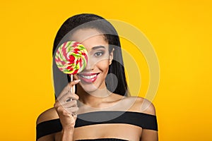 Smiling cute mulatto girl holding and covering her eye with big colourful sweet lollipop looking at camera over sunny
