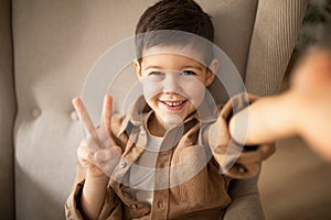 Smiling cute small european boy sitting on armchair, makes selfie and peace sign with hand in room interior