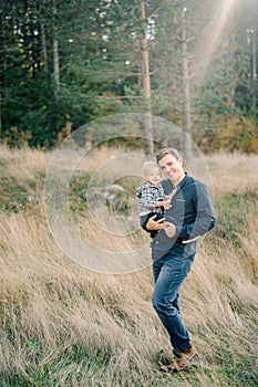 Smiling dad with a little daughter in his arms stands in dry grass at the edge of the forest