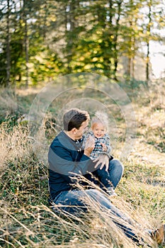 Smiling dad with a little girl on his knees sit on the lawn in the park