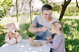 Smiling dad treating little girl with tea from a toy cup at a table in the garden having a tea party