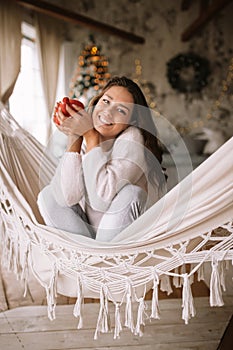 Smiling dark-haired girl dressed in pants, sweater and warm slippers holds a red cup sitting in a hammock in a cozy
