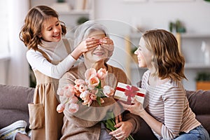 Smiling daughter and granddaughter giving flowers to grandmother celebrate spring holiday Women's Day at home