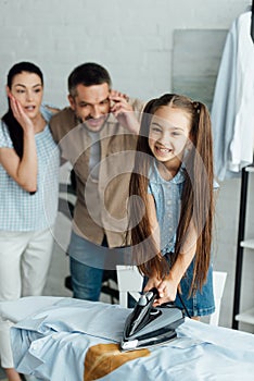 smiling daughter holding iron above burned shirt at home