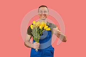 Smiling delivery man with bouquet of yellow flowers shows thumbs up on pink background. Flowers delivery concept