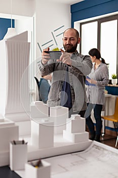 Smiling design architect holding smartphone to photograph desk with architectural model