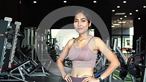 Smiling and determined youthful Asian female athlete looking at camera while putting hands on hips and standing at gym