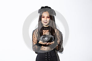 Smiling devious asian girl in witch costume, celebrating halloween, holding black pumpkin, standing over white