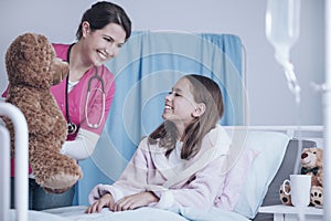 Smiling doctor in pink uniform giving plush toy to happy sick ch