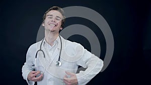 Smiling Doctor showing thumbs up, standing on a black background putting on a stethoscope. Packshot.