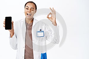 Smiling doctor shows okay sign, mobile phone screen app, medical clinic application, stands over white background