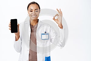 Smiling doctor shows okay sign, mobile phone screen app, medical clinic application, stands over white background