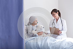 Smiling doctor with stethoscope talking with kid with cancer hugging plush toy