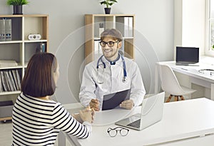 Smiling doctor talking to patient sitting at desk in medical office, clinic or hospital