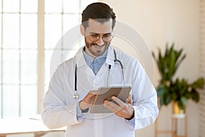 Smiling doctor wearing glasses using tablet, standing in office