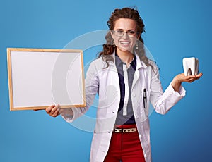 Smiling doctor woman showing tooth and blank board on blue