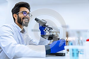 Smiling doctor working in modern laboratory research scientist examines substance under microscope.