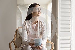 Smiling dreamy young woman holding cup, sitting in cozy armchair