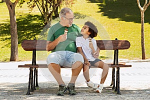 Smiling elderly european grandfather and mixed race little boy eat ice cream in park on bench