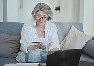 Smiling elderly female bank client making quick online payments with her credit card and laptop