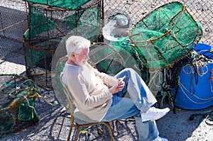 A smiling elderly fisherman on the dock of the harbor relaxes after work using his mobile phone, sitting near the fishing nets to