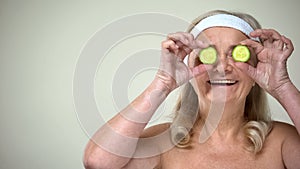 Smiling elderly lady closing eyes with slices of cucumber, skincare procedures