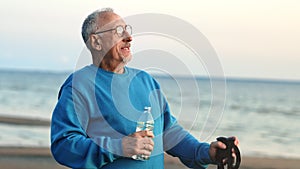 Smiling elderly man Scandinavian walking stick drinking water bottle contemplate sea sunset beach