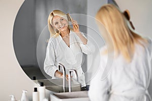 Smiling elderly woman in bathrobe combing hair while looking in bathroom mirror