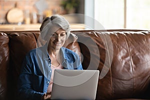 Smiling elderly woman in glasses using laptop at home