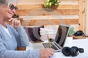 Smiling elderly woman gray hair working on laptop outdoor on a white table. Alternative outdoor office. Wooden background. Devices
