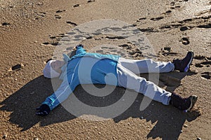 Smiling elderly woman lying on the sand in a star pose wearing a jacket and hat during the winter season. Active life in adulthood