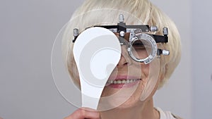 Smiling elderly woman wearing phoropter closing one eye, vision examination