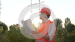 Smiling electrical engineer in helmet working on repair of electrical high voltage pylons outdoors, holding drawing