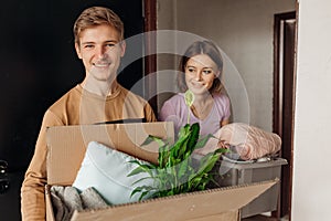 Smiling energetic students teenagers man, woman hold carton box with flower and clothes. Changeover new university room