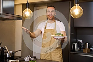 Smiling European man in beige chef's apron, holding a plate with Italian spaghetti pasta and showing it at camera