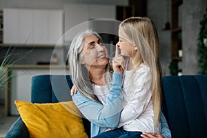 Smiling european old grandmother hugs small granddaughter, touches nose, enjoy free time