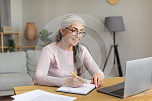 Smiling european old woman with gray hair in glasses look at computer, make notes at table