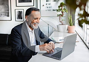 Smiling european senior businessman in suit typing on laptop at table in modern office interior