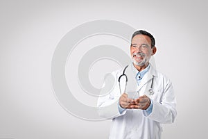 Smiling european senior doctor in white coat with stethoscope chatting on phone, looking at empty space