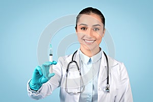 Smiling european woman doctor wearing sterile gloves, holding medical syringe, blue studio background