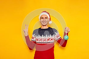 smiling, excited man in Christmas sweater with reindeer, red Santa Claus hat holds Christmas tree toy in his hand and