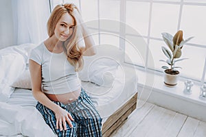 Smiling expectant lady sitting on edge of bed at home