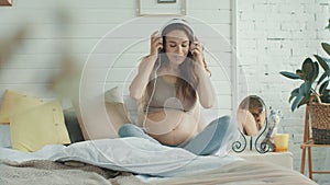 Smiling expectant mother wearing headphones on naked belly in bedroom.