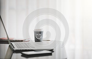 Smiling face on paper cup of hot coffee with laptop on desk besides window