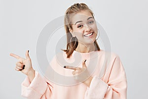 Smiling fair-haired young woman in pink sweatshirt pointing at copy space for your advertisment or text. Positive girl photo