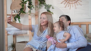 Smiling family in bed, where dad and mom play with their little daughter, mom takes a selfie of her family.