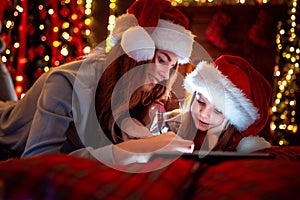 Smiling family mother and daughter in santas hats and pajamas watching funny video or choosing gifts on digital tablet