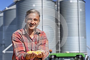 Smiling Farmer Showing Freshly Harvested Corn Maize Grains in front of Farm Grain Bins