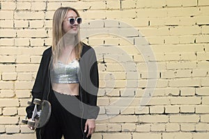 Smiling fashion woman with a skateboard on brickwall background. Girl in sunglasses with longboard